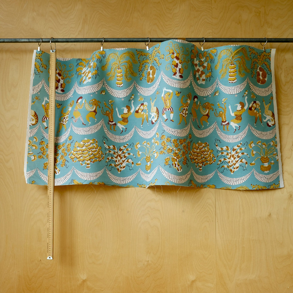 A piece of patterned fabric hanging from a metal rod against a light wood wall, with a measuring tape hanging alongside it for scale. The fabric has a turquoise blue background with a repeating pattern of gold and white decorative motifs.