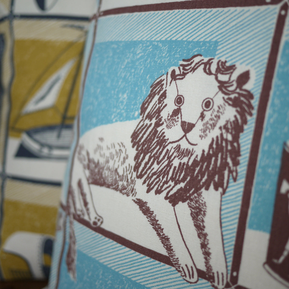 A close-up of a printed fabric cushion or pillow featuring a stylized lion illustration in dark brown on a light blue and white background, with a blurred matching patterned textile behind it.
