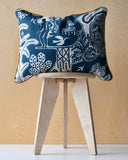 Decorative cushion with dark blue patterns and ochre trim on a wooden stool against a beige wall.
