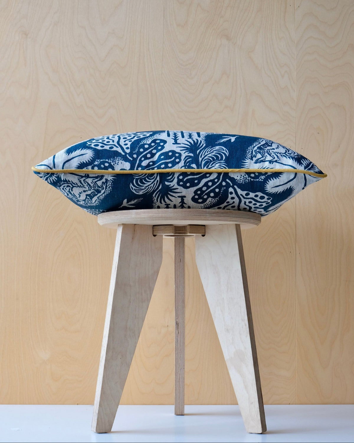 Blue patterned cushion with ochre trim on a wooden stool against a light wood panel background