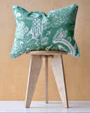 Green patterned cushion with pale blue trim on a wooden stool against a plywood wall. 