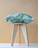 Green and white patterned cushion with pink trim on a wooden stool against a light wood panel background