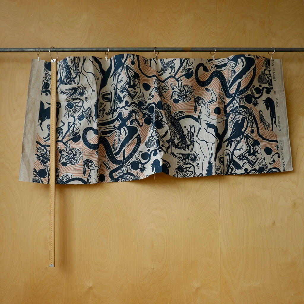 A patterned fabric panel or curtain hanging from a metal rod against a light wood wall, with a wooden measuring stick placed beside it for scale. The fabric has an abstract black and navy print over a warm beige/cream base with some peach-tan linework.